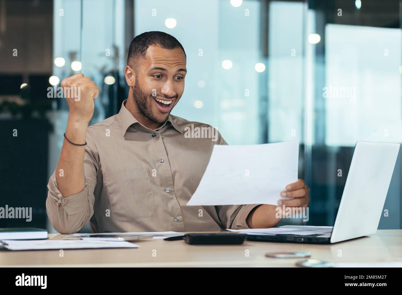 Portrait d'un homme d'affaires hispanique à succès, d'un homme en chemise lisant un rapport et heureux célébrant le succès satisfait du résultat tenant la main vers le haut, employé au bureau avec ordinateur portable travaillant sur papier. Banque D'Images