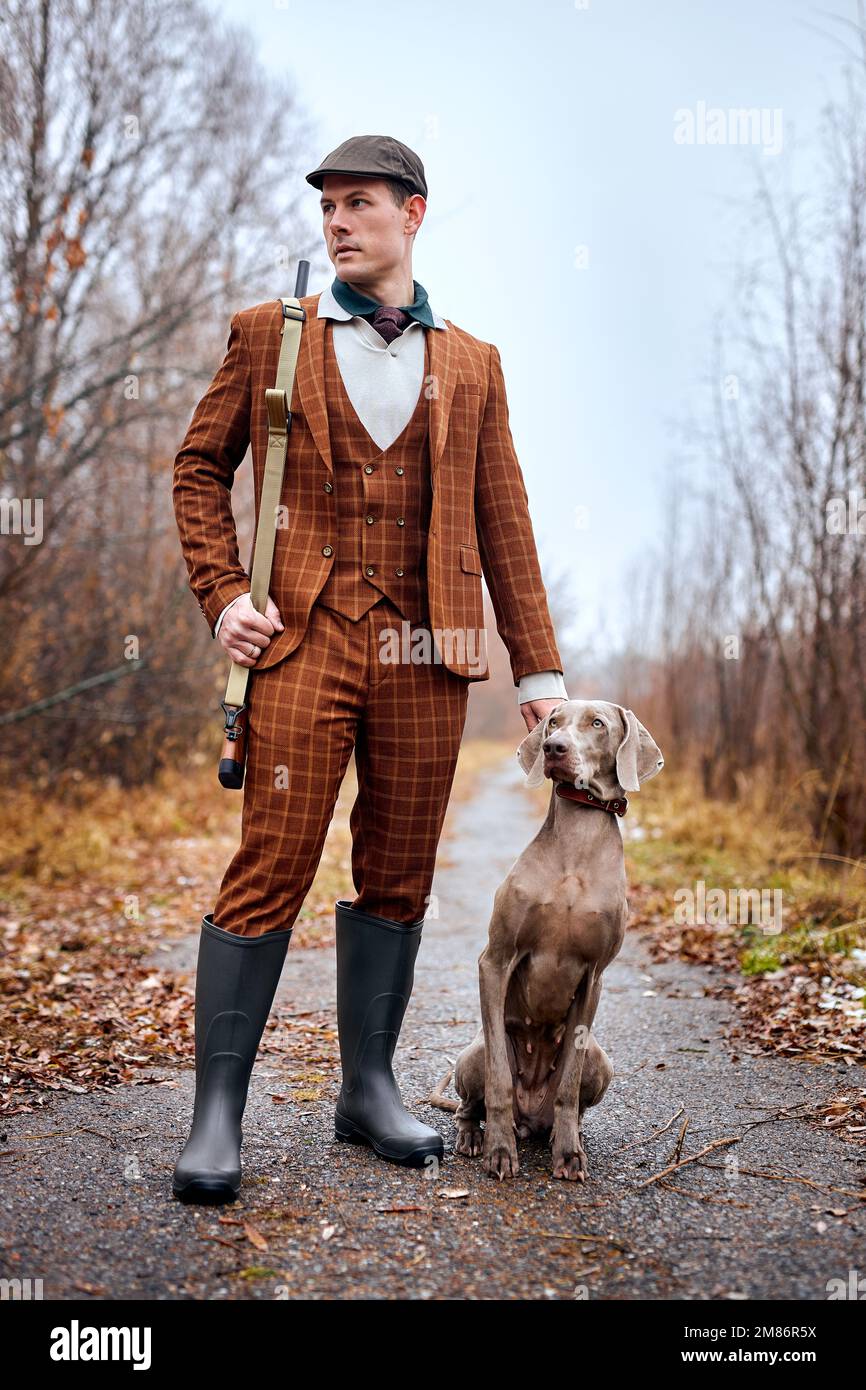 Jeune homme caucasien dans le chasseur de costume dans la campagne, homme tenant une arme et marchant avec le chien. Chasseur avec fusil de chasse. Chien très beau et fort waïtine Banque D'Images