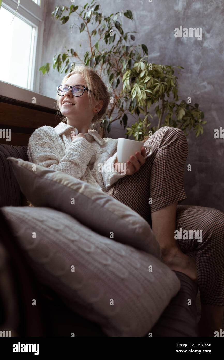 Verticale moderne, souriante, pensive et confortable blonde pieds nus femme en lunettes s'assoir sur un canapé avec coussins et boire du thé chaud Banque D'Images