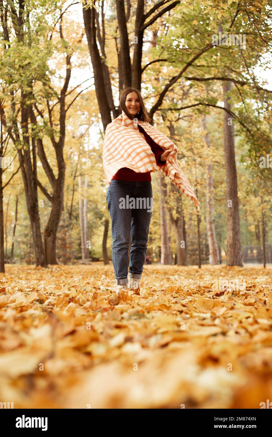 Femme souriante et timide, enveloppée d'un foulard à carreaux chaud et d'un pull, se penche sur le tronc de l'arbre, près de la zone des feuilles dorées Banque D'Images