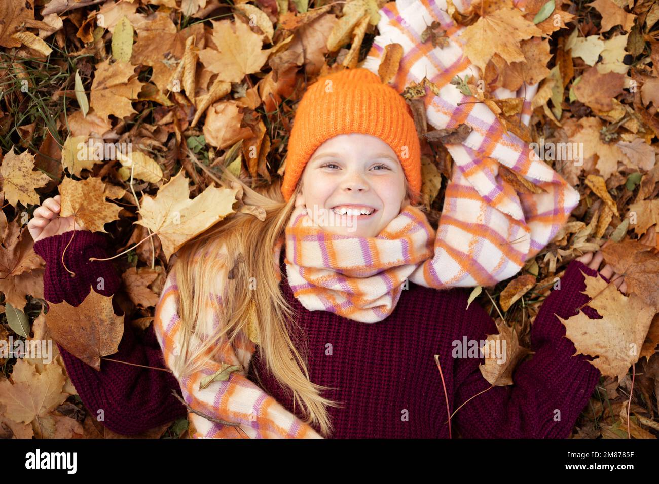 Positif heureux et souriant, ravissante petite fille blonde dans un ensemble chaud couché, tenir des feuilles colorées sur le feuillage d'automne Banque D'Images