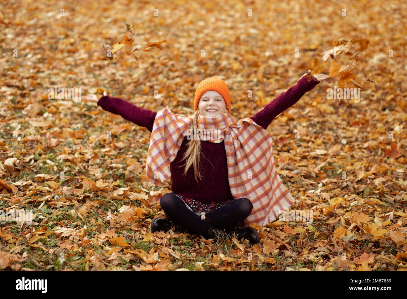 Rire actif positif joueur petite fille blonde en orange chaud chapeau, écharpe, pull assis, jeter des feuilles sur le terrain d'automne Banque D'Images