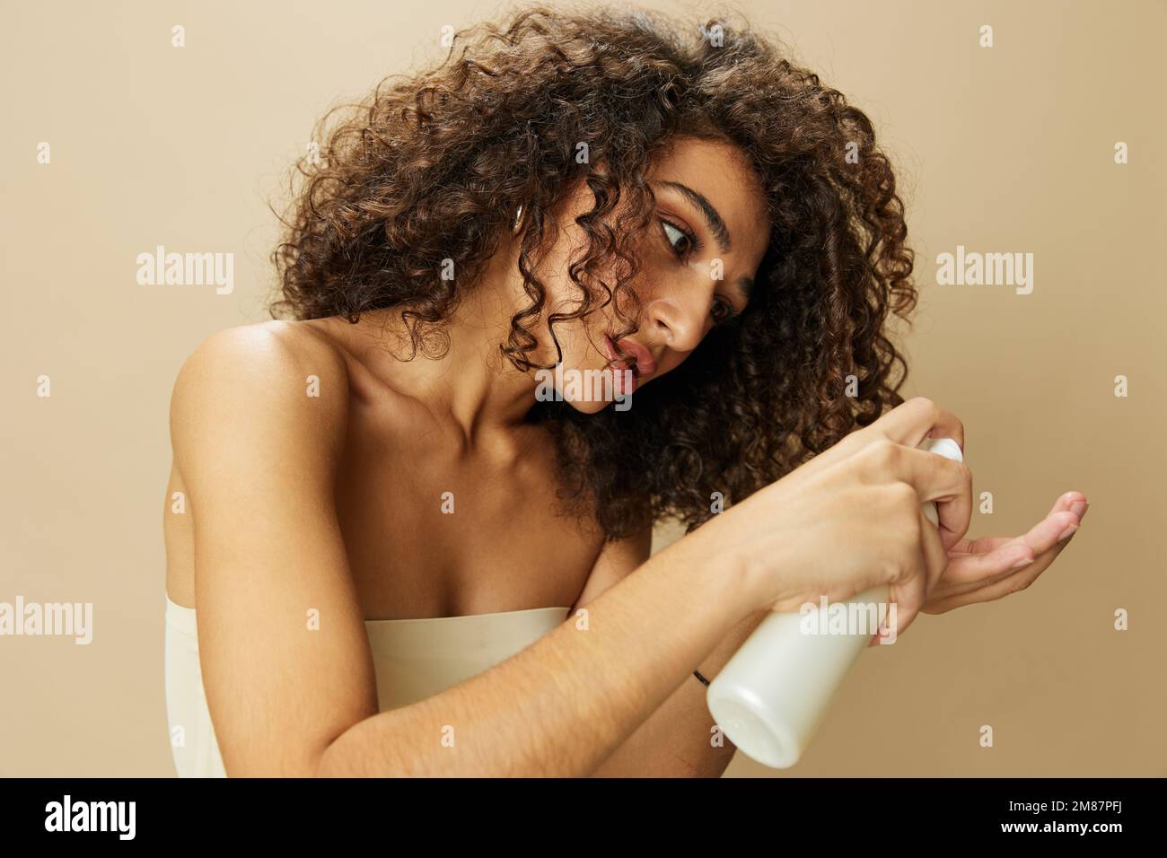 Femme applique de la crème et du baume à ses cheveux bouclés, le concept de protection et de soin avec des produits de salon, un look sain, un sourire aux dents sur un beige Banque D'Images