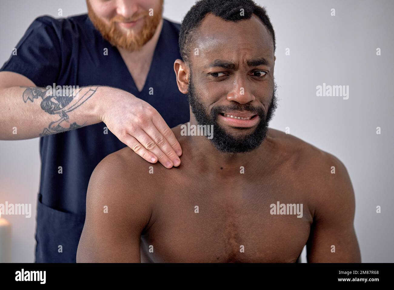 Un thérapeute caucasien court fait un massage relaxant pour un homme noir d'âge moyen, le frottant contre le dos. Un homme barbu frustré qui se guérit de la colonne vertébrale Banque D'Images