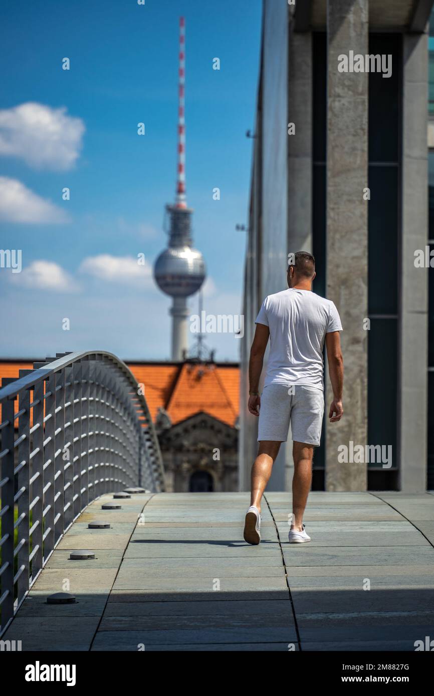 Un jeune homme blanc traverse Marie-Elisabeth-Lüders-Steg, un pont public piétonnier, avec la célèbre tour de télévision Alexanderplatz au loin Banque D'Images