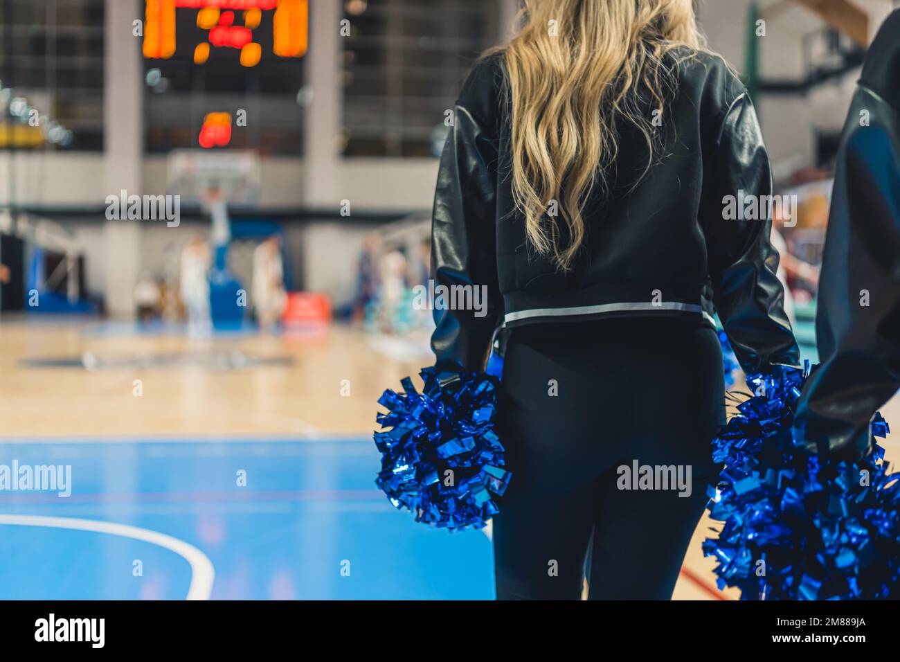 Un meneur de l'arrière dans un stade pendant un match. Elle tient des pompons dans ses mains. La fille est vêtue d'une veste noire et d'un pantalon ajusté. Les joueurs peuvent être vus en arrière-plan. Banque D'Images