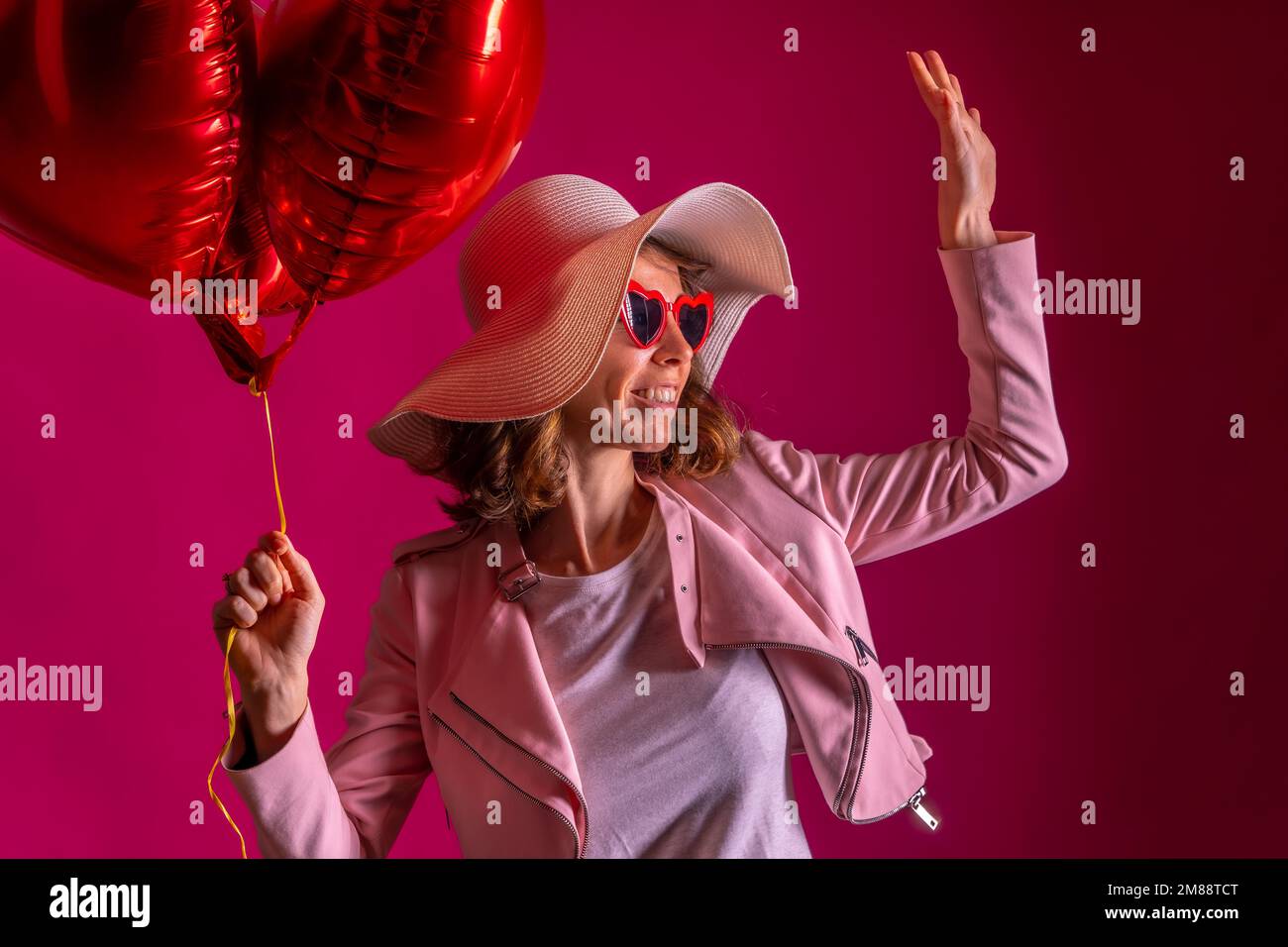 Une femme caucasienne avec un chapeau blanc dans une boîte de nuit avec quelques ballons de coeur, fond rose Banque D'Images