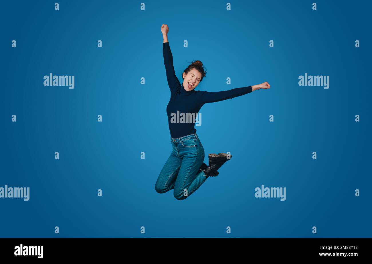 Une femme excitée et surjoyeuse se porte en sautant haut avec des mains tendues isolées sur un portrait de studio à fond bleu. Concept de mode de vie des personnes Banque D'Images