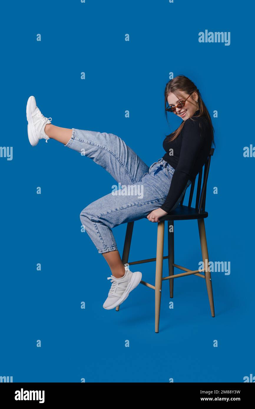 Photo d'une charmante femme portant des lunettes assise sur une chaise levant les jambes isolées sur fond bleu. Banque D'Images