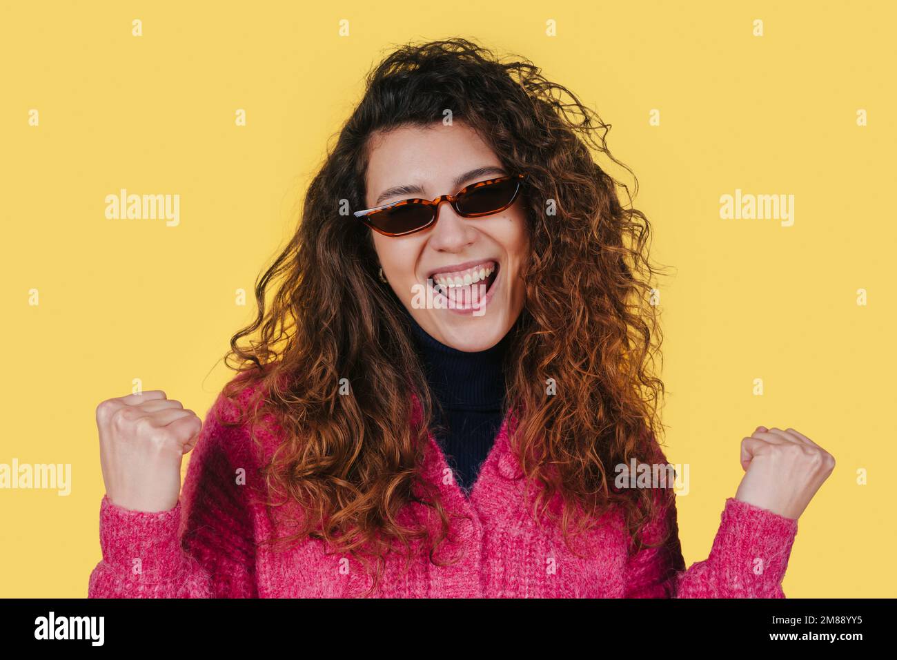 Photo d'une femme gaie avec des cheveux bouclés clenches Fists célébrant le succès portant des lunettes de soleil isolées sur fond jaune. Les gens et le bonheur Banque D'Images
