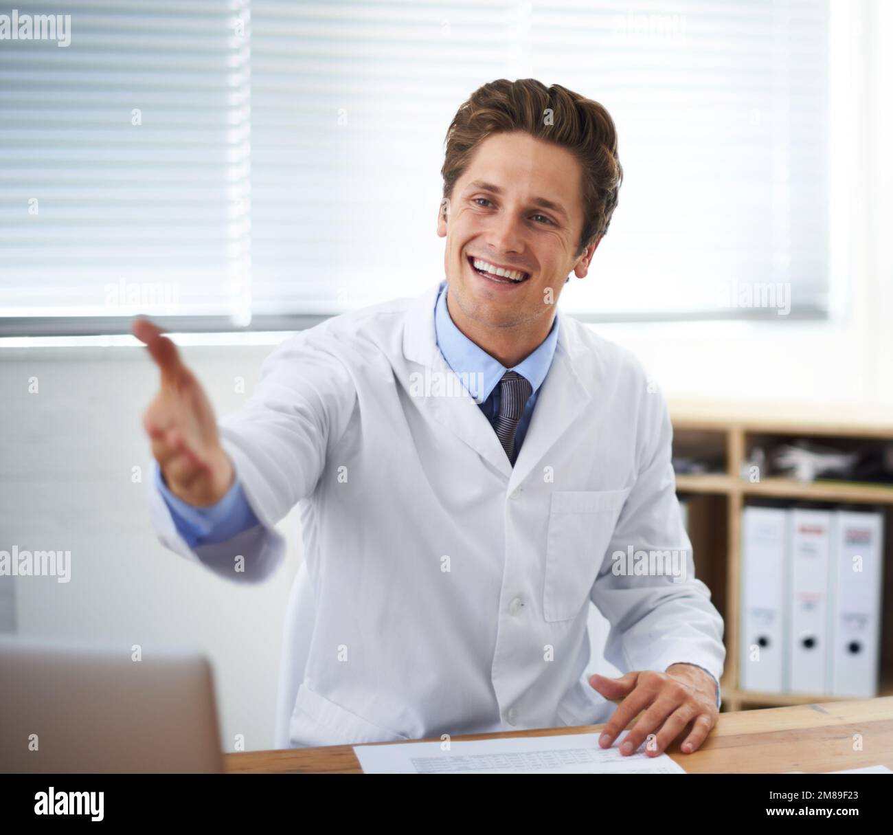 Je vous réjouis de pouvoir prendre rendez-vous. Portrait d'un jeune médecin à l'aspect positif assis dans son bureau et faisant un geste pour Entrez. Banque D'Images
