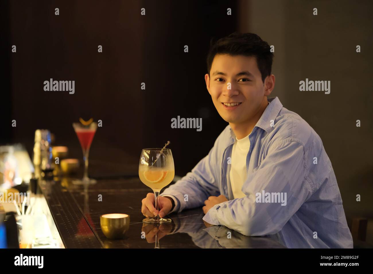 Un jeune homme asiatique s'assoit au comptoir du bar avec un verre de cocktail, sourire à la caméra Banque D'Images