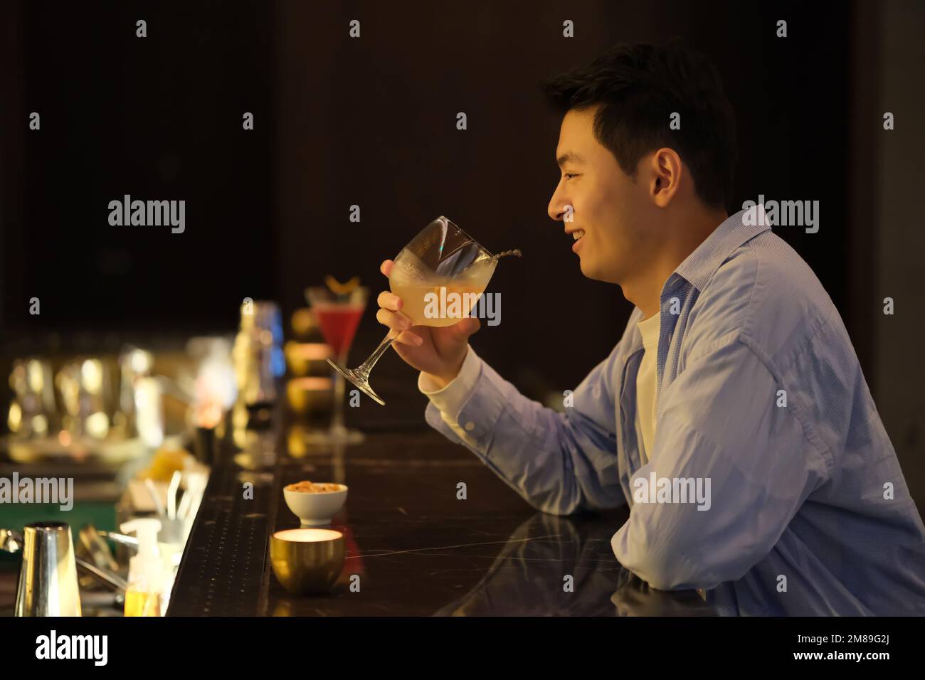 Un jeune asiatique tient et boit un verre de cocktail au comptoir du bar, vue latérale Banque D'Images