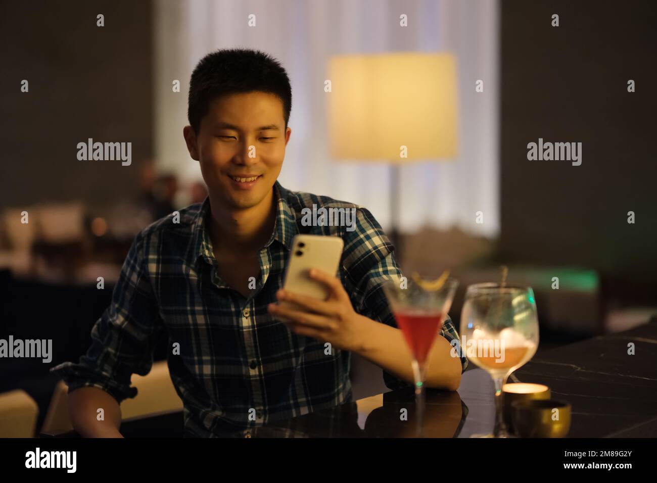 Un jeune homme asiatique souriant regarde le téléphone au comptoir du bar de nuit Banque D'Images