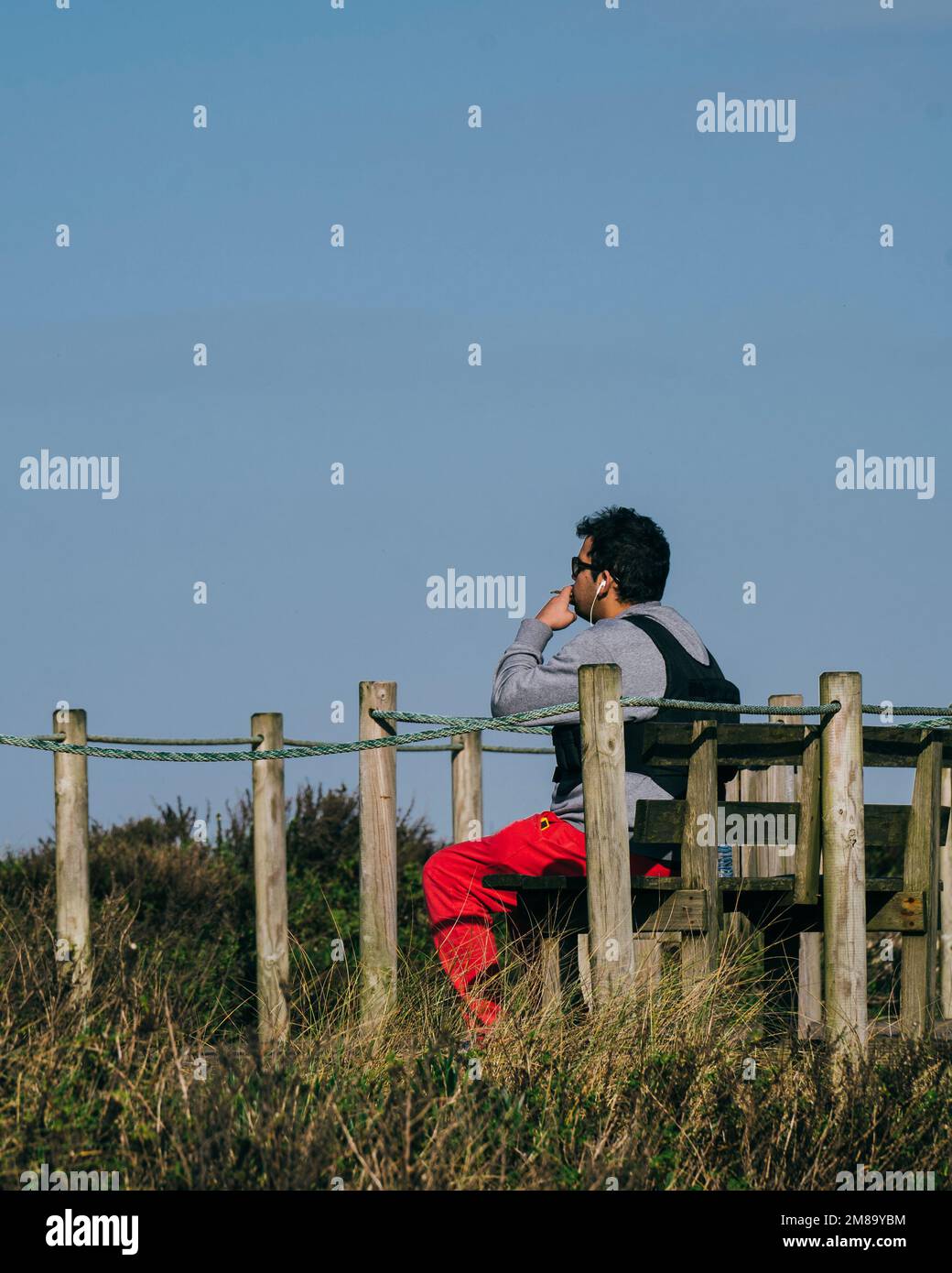 Un jeune homme fume et se détend sur un banc en bois qui donne sur la mer tout en écoutant de la musique Banque D'Images