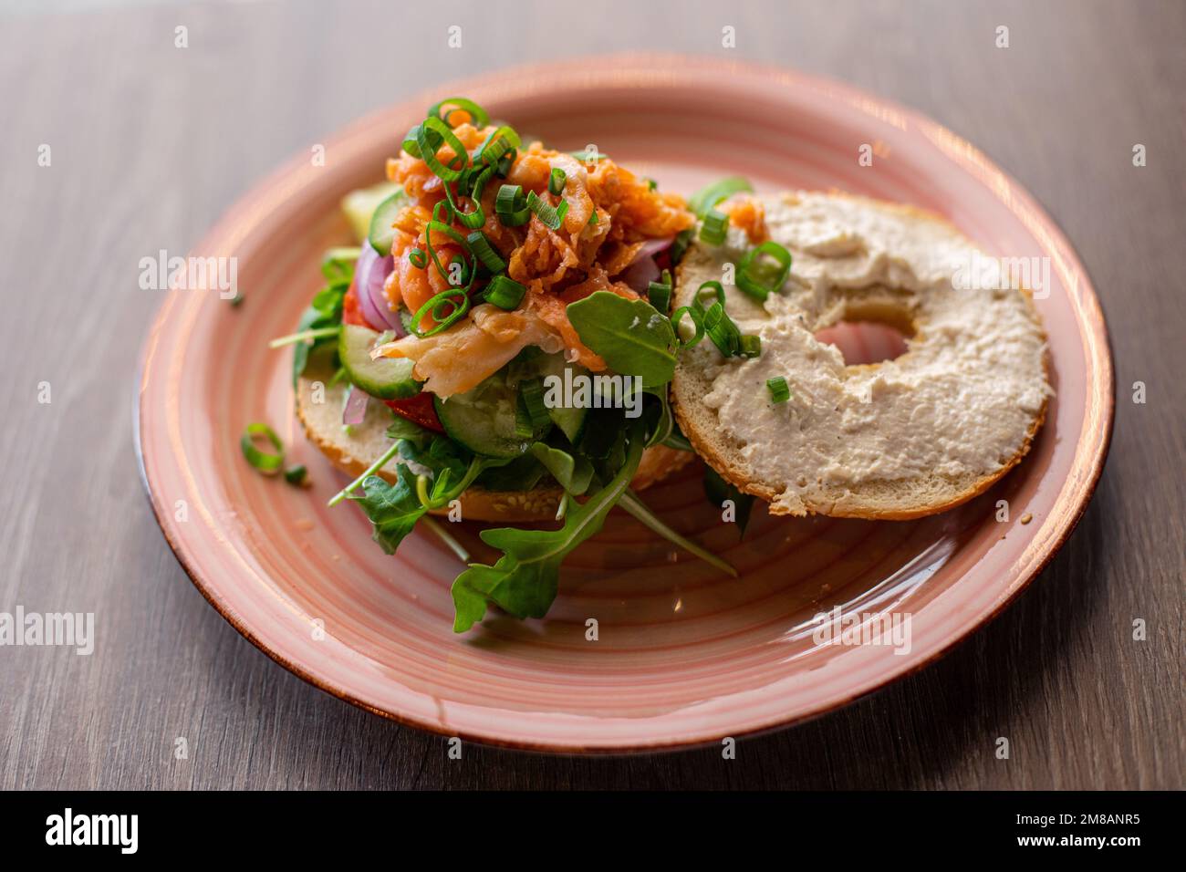 Vue latérale d'une assiette rose avec hamburger à dents ouvertes avec trou dans un petit pain, saumon, arugula, verdure, oignon vert, concombres. Banque D'Images Vue latérale d'une assiette rose avec hamburger à dents ouvertes avec trou dans un petit pain, saumon, arugula, verdure, oignon vert, concombres. Banque D'Images