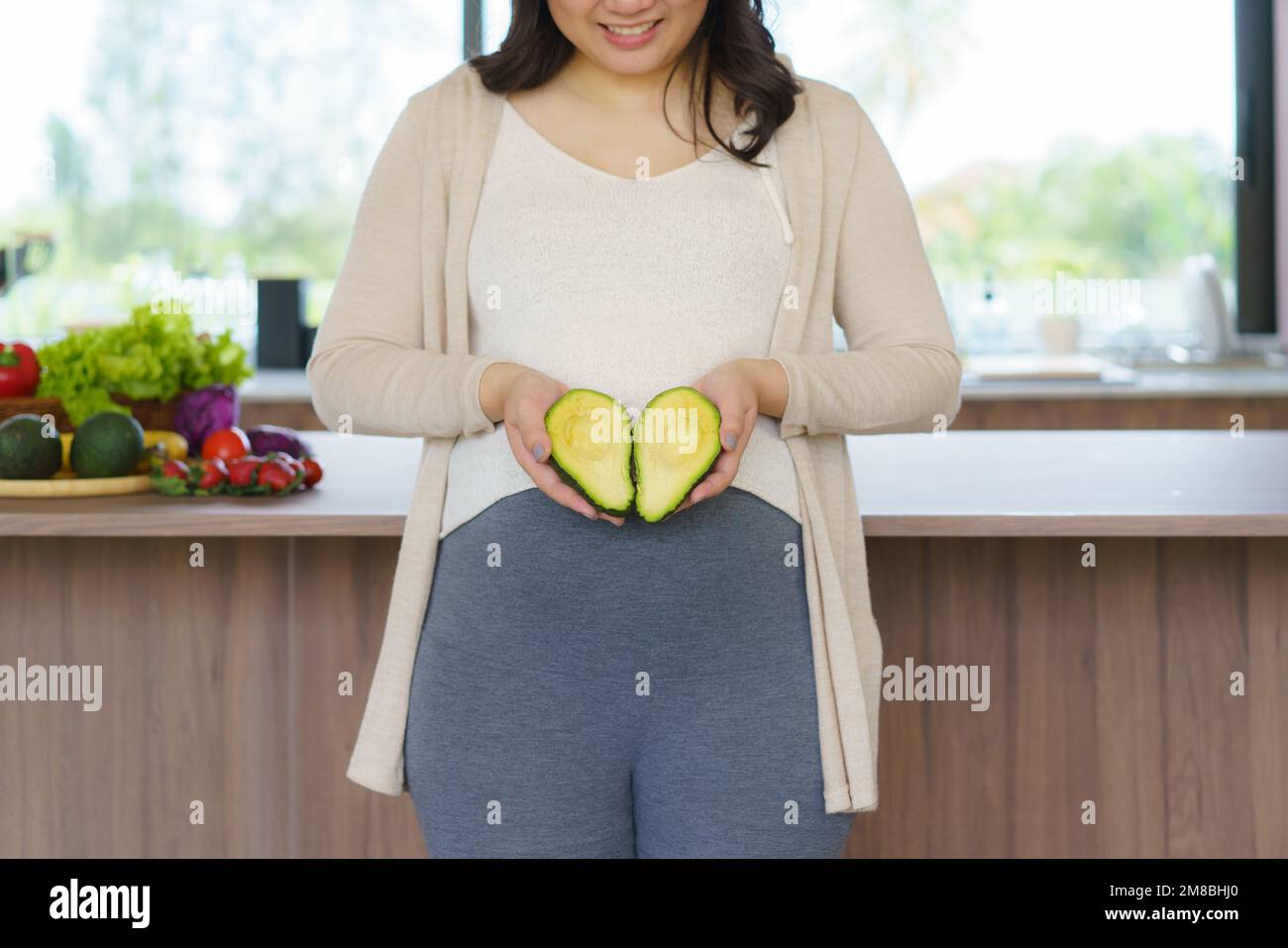Femme enceinte asiatique tenant des fruits frais d'avocat près de son ventre dans la cuisine à la maison. Nutrition saine et concept de grossesse Banque D'Images