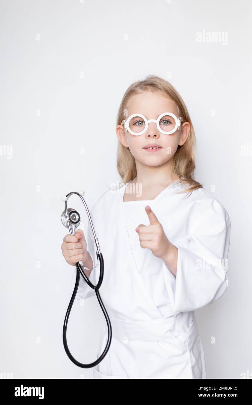 Petite fille sérieuse vêtue dans une tenue de docteur avec des lunettes de jouet, tenant le stéthoscope, pointant avec l'index vers le haut dans le mouvement d'arrêt ou d'interdiction. Bas Banque D'Images