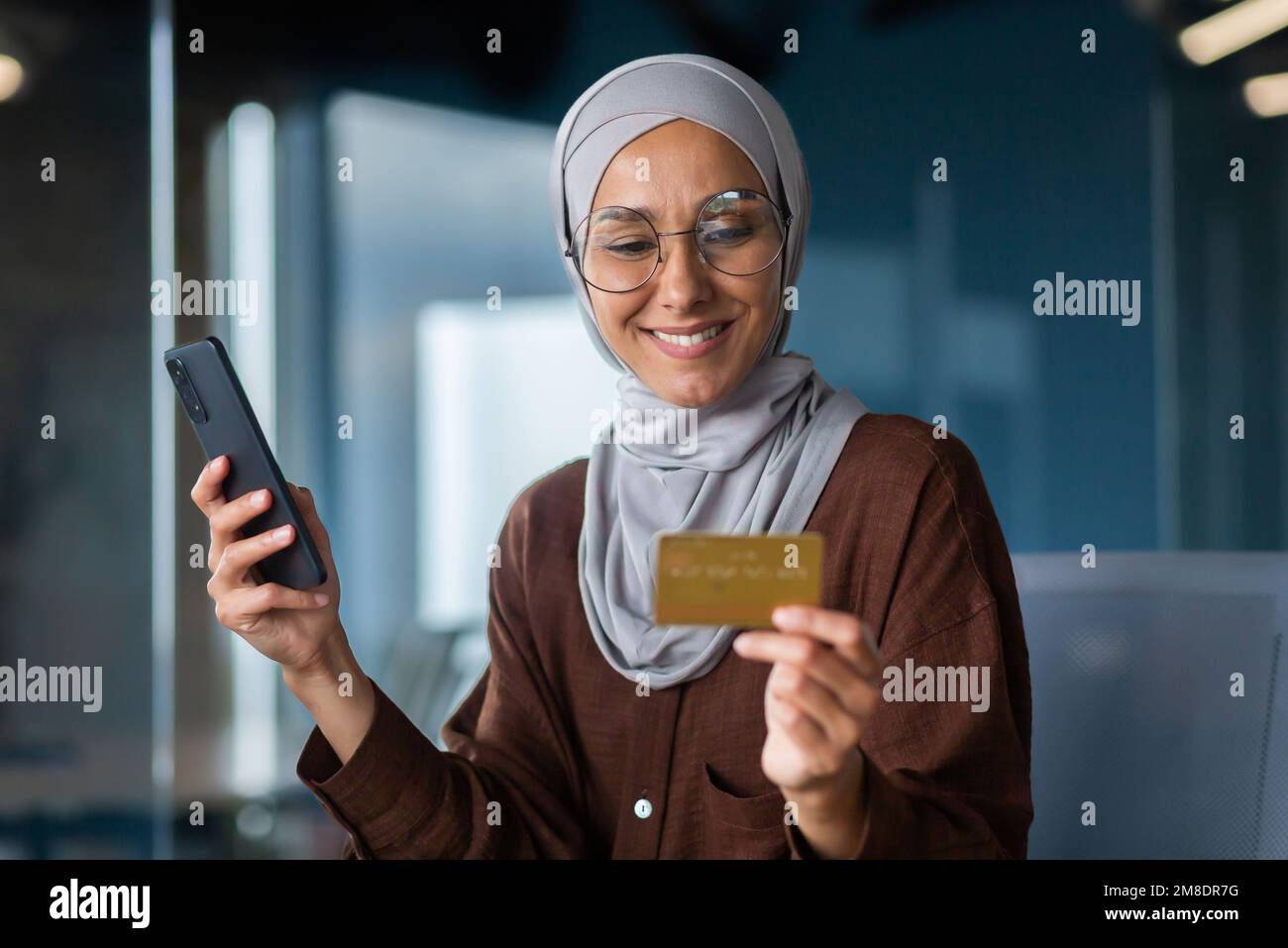 Femme d'affaires prospère dans le hijab travaillant à l'intérieur du bureau avec un ordinateur portable sur le lieu de travail, femme musulmane tenant le téléphone et la carte de crédit bancaire pour les achats en ligne et le transfert d'argent. Banque D'Images