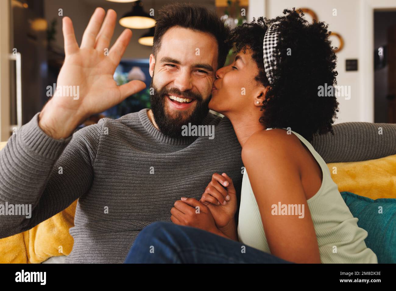Heureux biracial barbu homme en agitant la main et petite amie l'embrassant sur la joue tout en se relaxant sur le canapé Banque D'Images