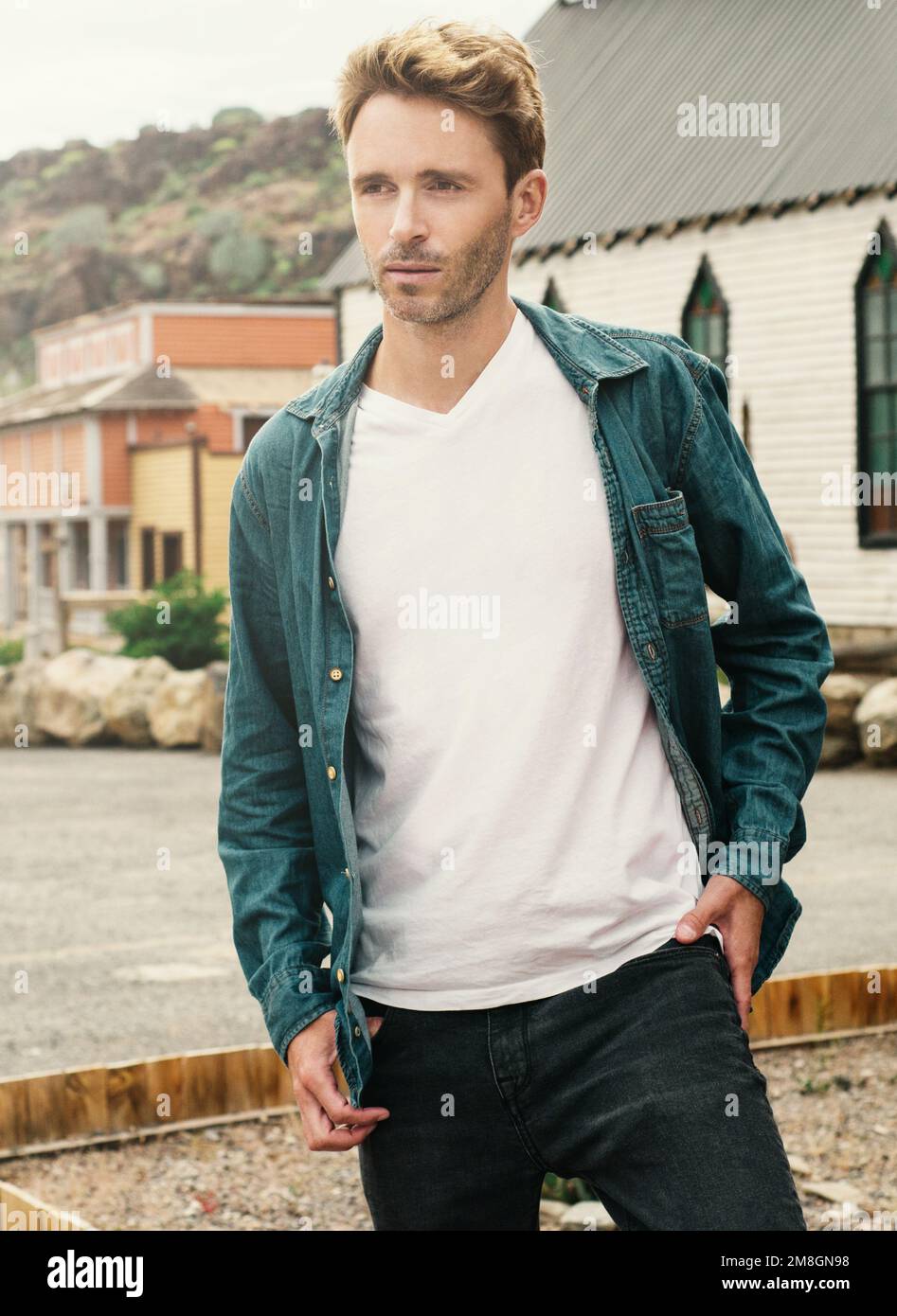 Un jeune homme beau est debout sur un fond de ville de l'Ouest. L'homme porte un t-shirt blanc vide sans logo. Maquette verticale. Banque D'Images