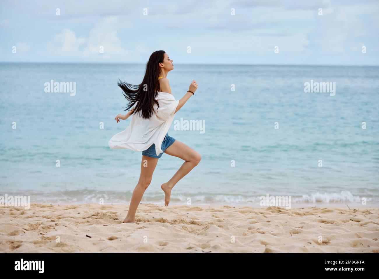 Sport femme court le long de la plage en vêtements d'été sur le sable dans un débardeur jaune et jeans short blanc shirt vol cheveux vue sur l'océan, plage Banque D'Images