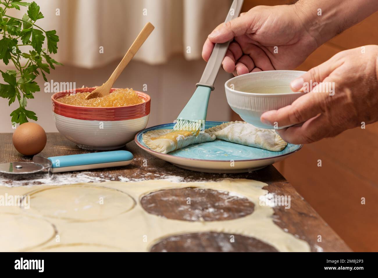 Les mains de l'homme avec un bol et une brosse de cuisine étalant les oeufs battus sur les bonbons de pâte feuilletée avant des mettre dans le four. Banque D'Images