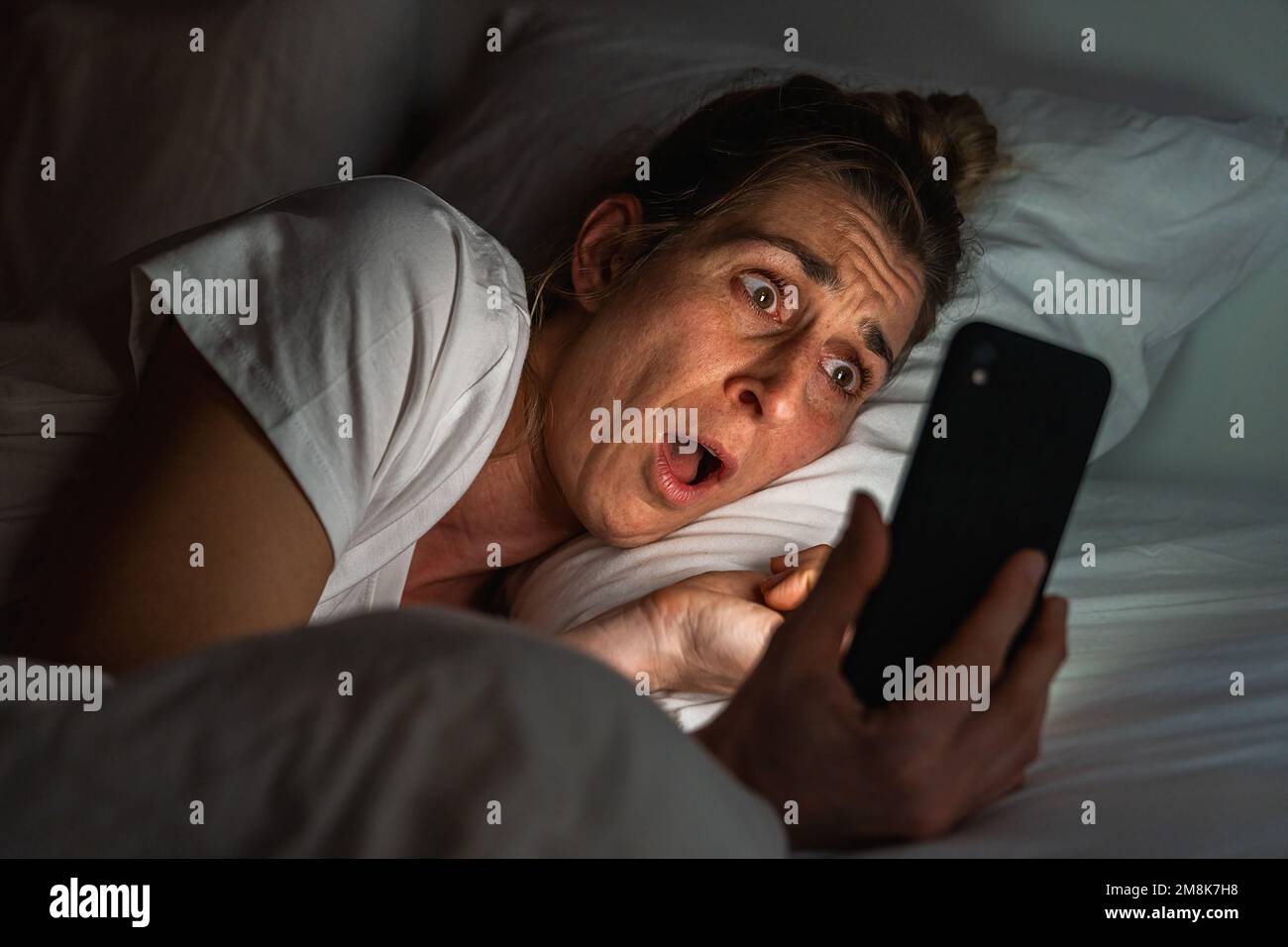 Fille surprise utilisant un smartphone dans le lit. L'insomnie et la dépendance des médias sociaux concept. Banque D'Images