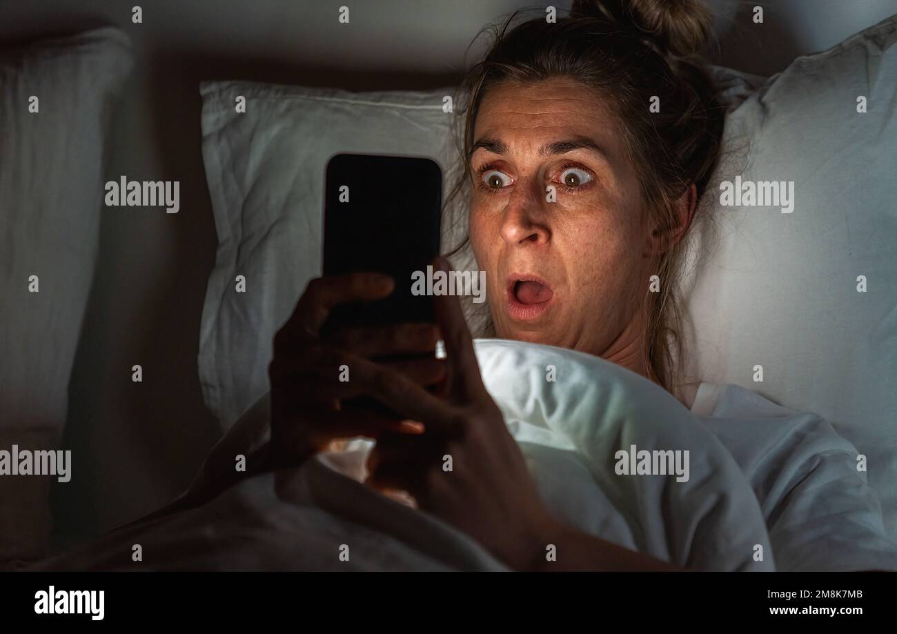 Femme choquée utilisant un smartphone dans le lit. L'insomnie et l'image de concept de harcèlement Banque D'Images