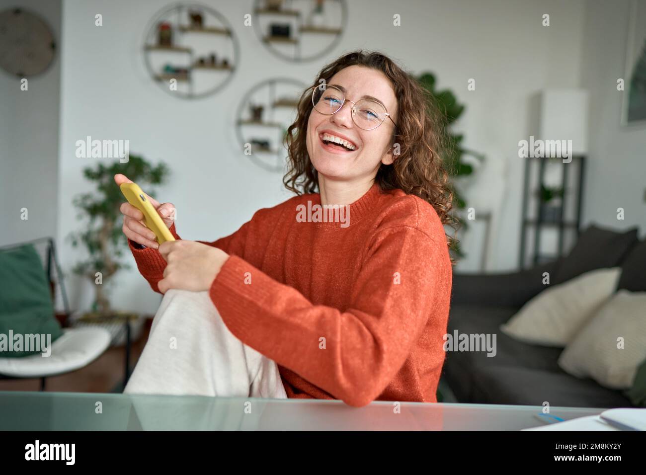 Bonne jolie jeune femme riant assis à la maison à l'aide d'un téléphone portable. Banque D'Images