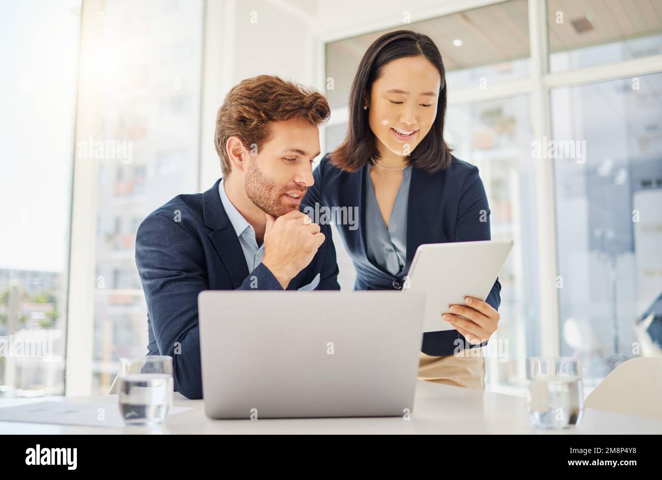 Deux jeunes collègues heureux et divers travaillant ensemble sur une tablette numérique dans un bureau. Femme d'affaires asiatique confiante montrant un caucasien souriant Banque D'Images