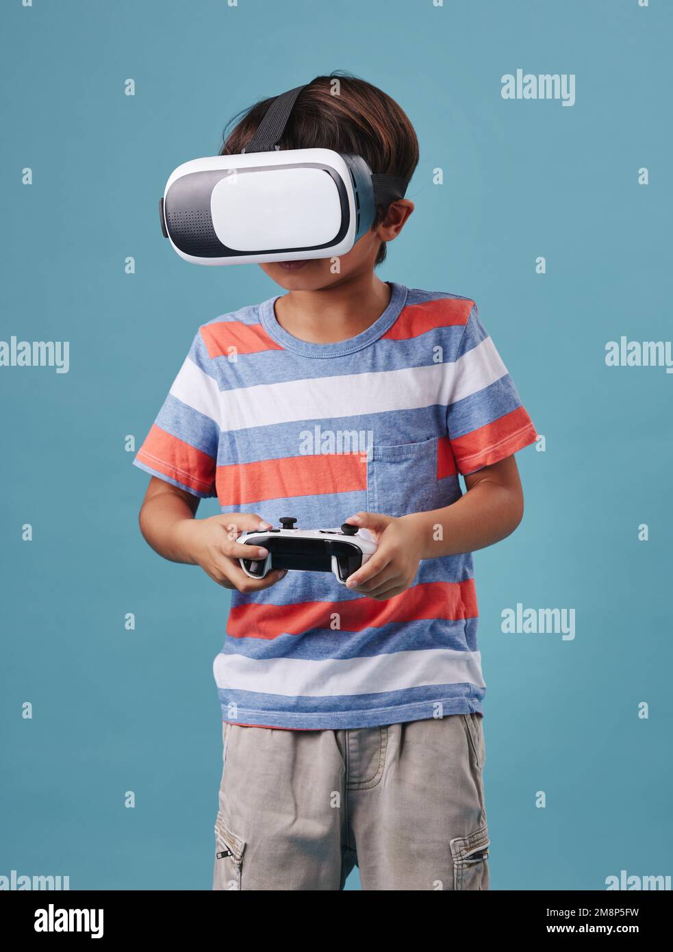 Jeune garçon de course mixte debout et portant un casque VR sans fil et tenant une manette de console tout en jouant à un jeu vidéo sur fond bleu Banque D'Images