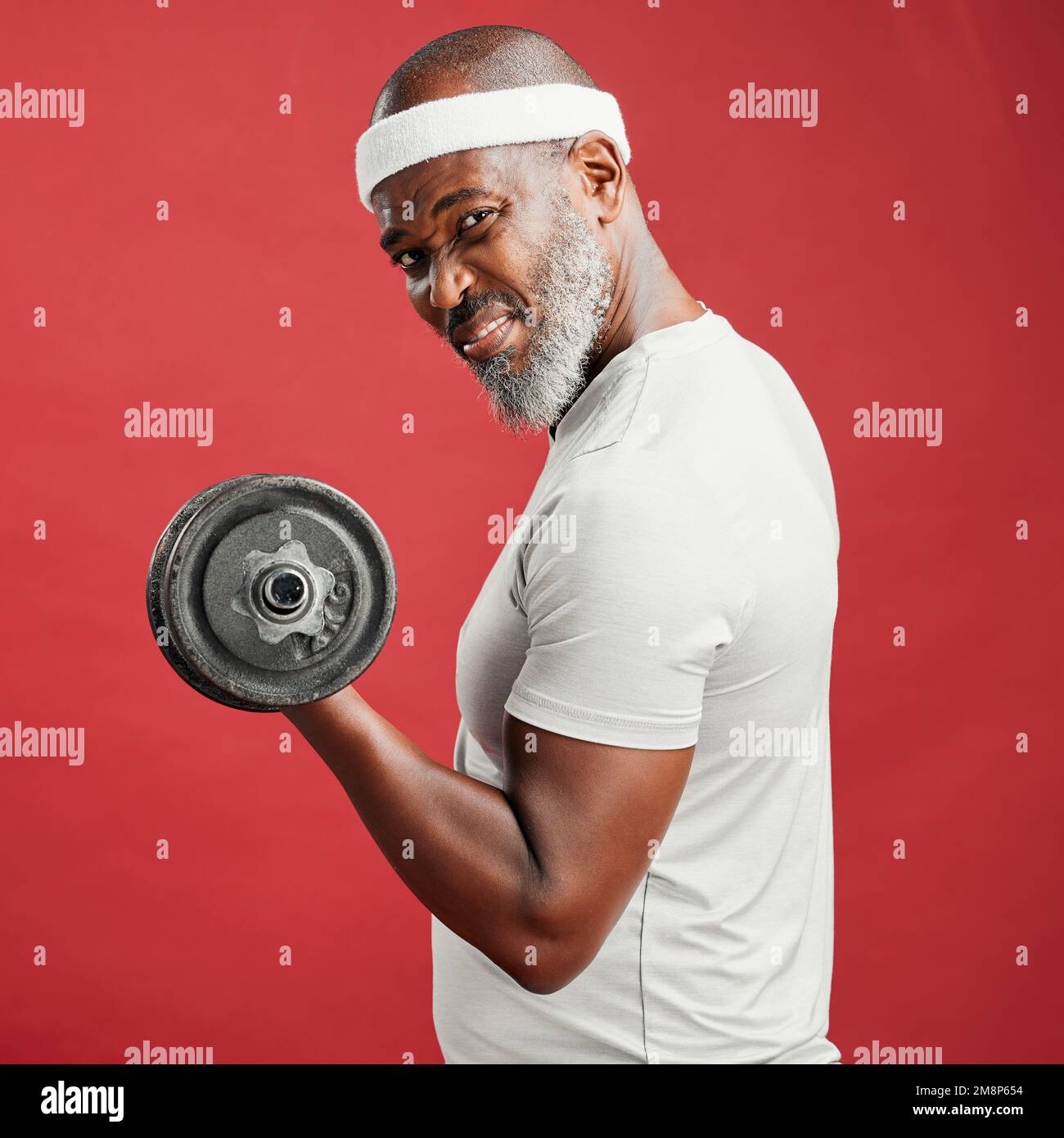 Homme afro-américain mature et confiant, debout seul sur fond rouge dans un studio et posant avec des haltères. Homme noir concentré et sportif Banque D'Images