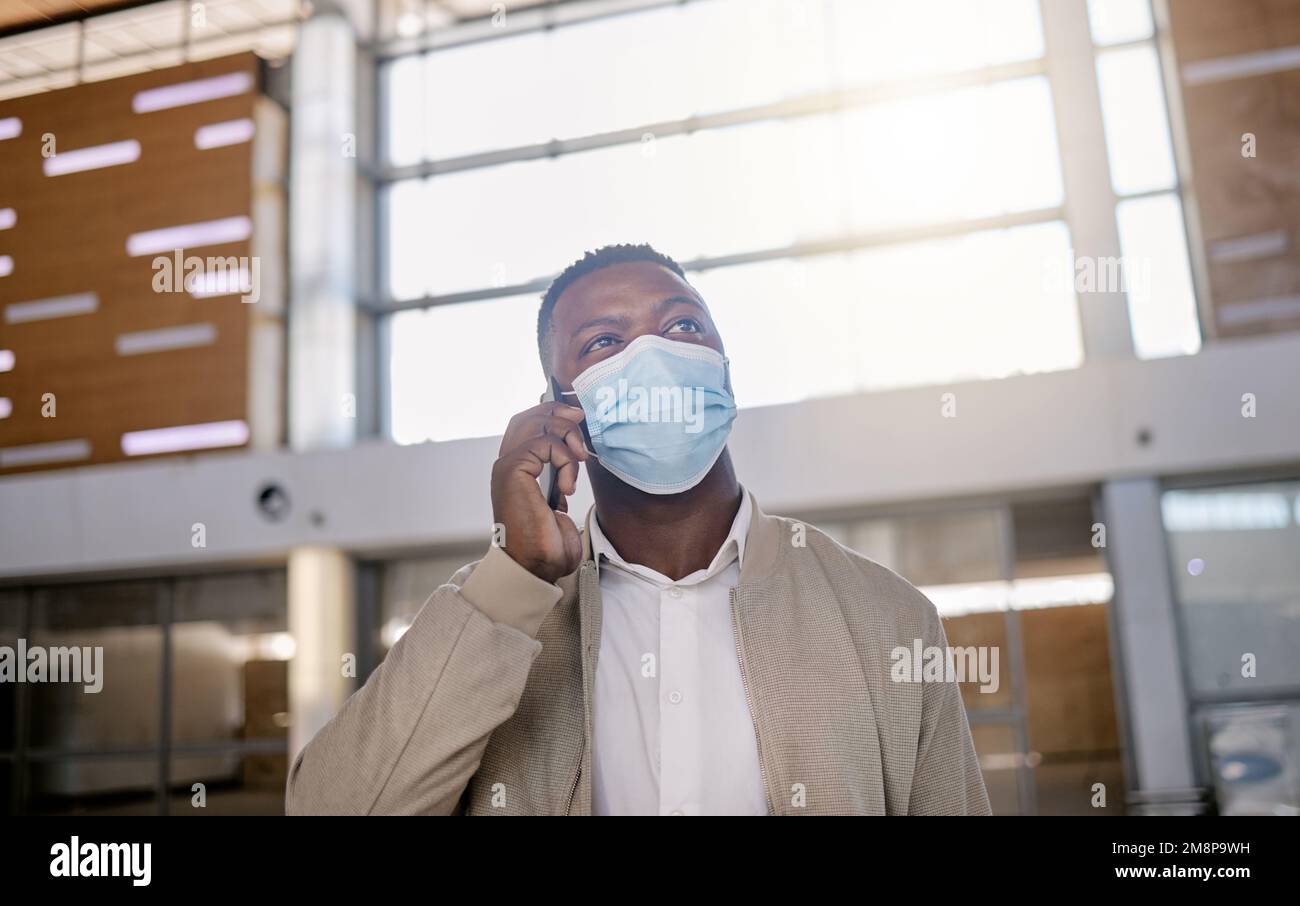 Un homme afro-américain est en communication téléphonique avec son appareil mobile à l'intérieur d'un poste pendant la journée, tout en portant un masque. Jeune homme noir parlant au téléphone Banque D'Images