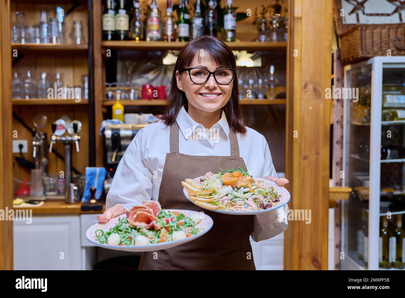 Femme d'âge moyen propriétaire du restaurant en tablier avec des assiettes de plats cuisinés Banque D'Images