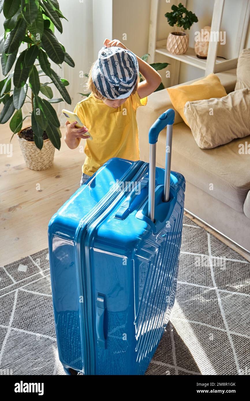 Enfant utilisant un téléphone portable achetant des billets en ligne ou attendant des vacances à la maison à côté d'une valise emballée Banque D'Images