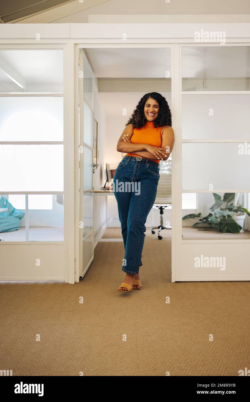 Femme entrepreneur réussie souriant à la caméra tout en se tenant dans un bureau avec ses bras croisés. Bonne jeune femme d'affaires travaillant dans un créativ Banque D'Images