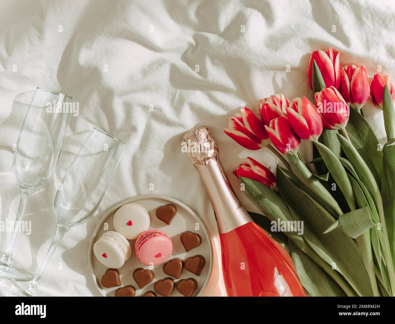 Bouquet de tulipes rouges, vin rose, verres à champagne, macarons et bonbons en forme de coeur sur draps blancs. Concept de la Saint-Valentin, date, mars 8. Fleurs de printemps et bouteille. Plan d'agencement avec vue de dessus. Copier l'espace Banque D'Images