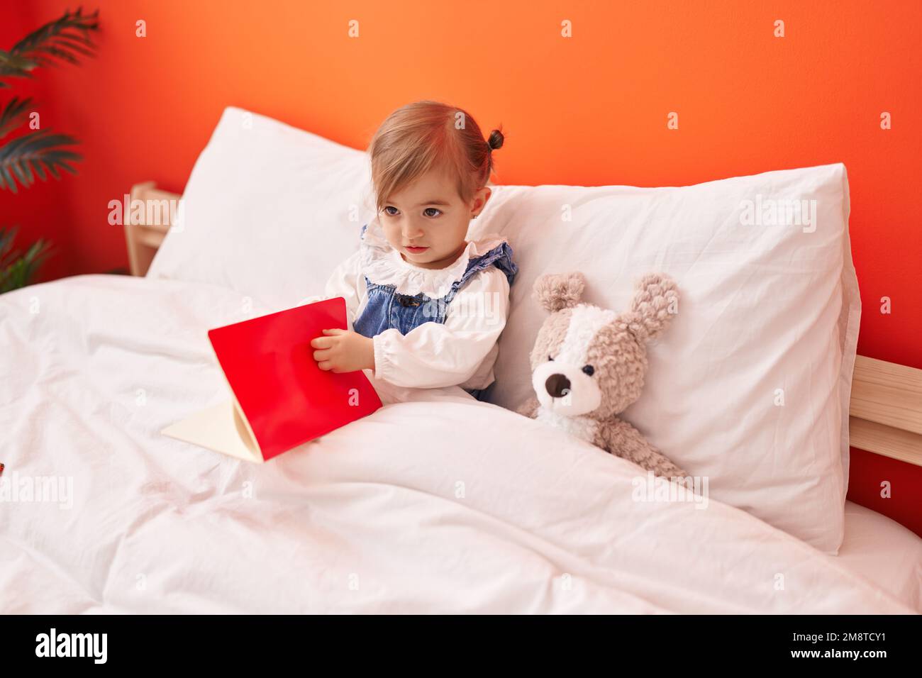Adorable livre de lecture blond pour tout-petit assis sur le lit avec un ours en peluche dans la chambre Banque D'Images