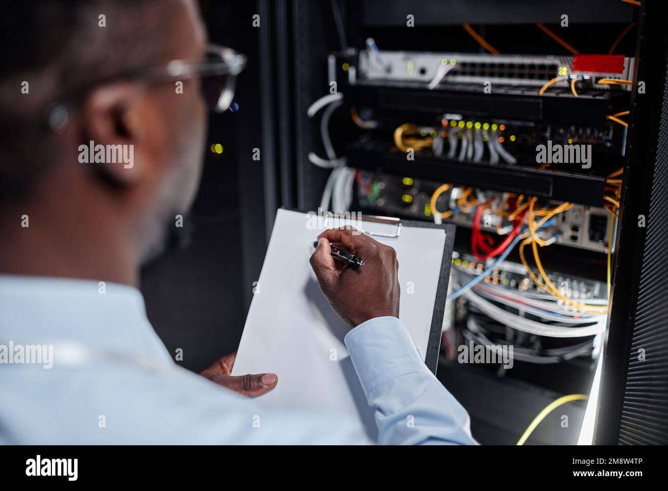 Gros plan de l'homme noir travaillant avec l'armoire de serveur dans le centre de données et prenant des notes sur le presse-papiers, l'espace de copie Banque D'Images