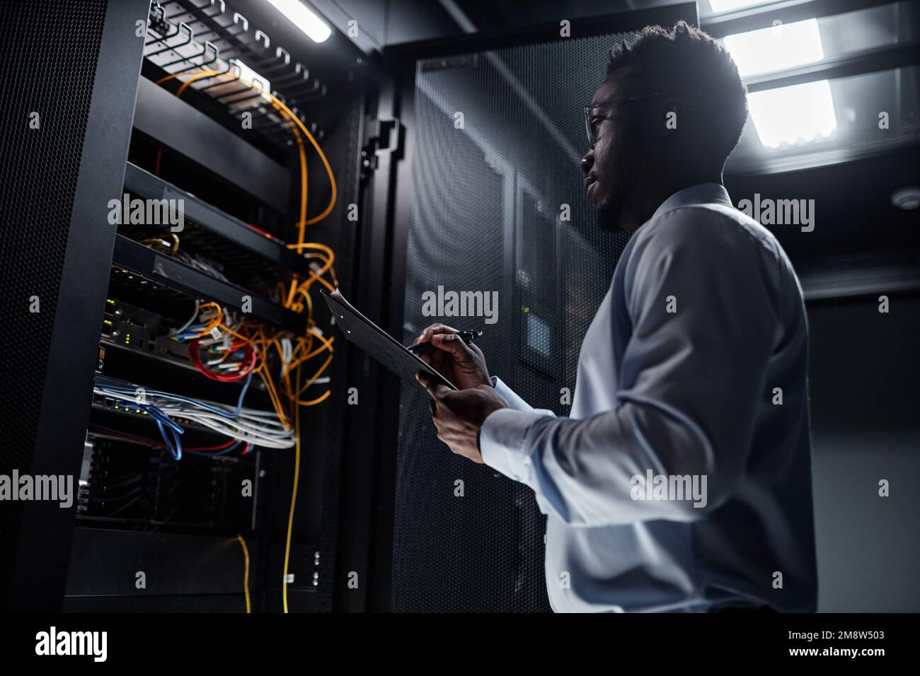 Portrait avec vue latérale rétroéclairé d'un homme noir travaillant avec l'armoire de serveur dans le centre de données et prenant des notes sur le presse-papiers, l'espace de copie Banque D'Images