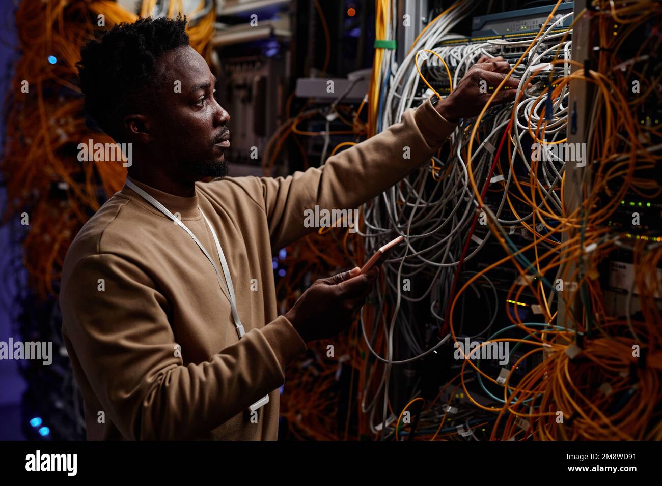 Vue latérale d'un jeune homme noir en tant qu'administrateur système qui fixe le serveur avec des câbles et des fils désordonnés Banque D'Images