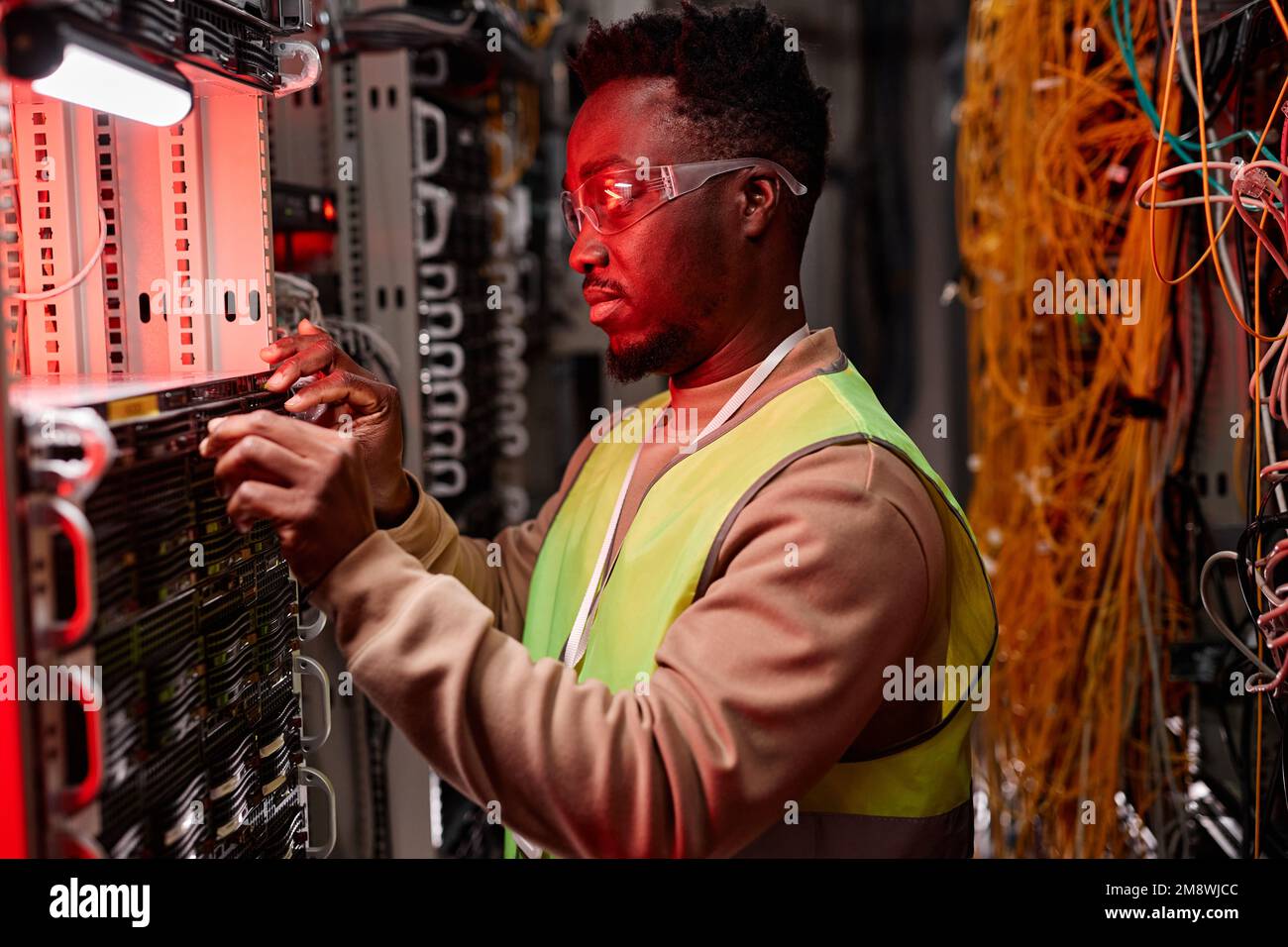 Vue latérale d'un technicien réseau inspectant les serveurs du centre de données, éclairé par une lumière néon rouge Banque D'Images