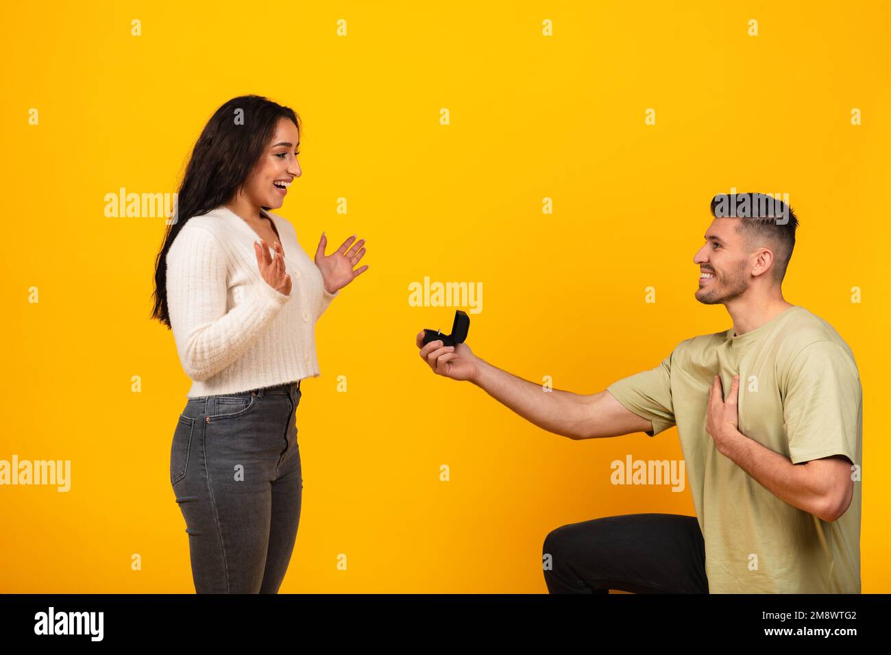 Souriant millennium arabe gars à genoux donnant boîte à anneau à la mariée heureuse excitée, isolée sur fond orange Banque D'Images