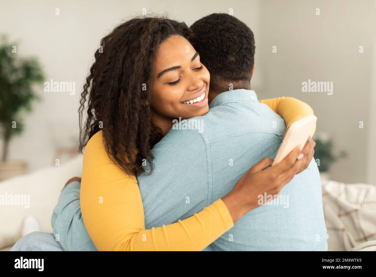Une jeune femme afro-américaine gaie qui embrasse l'homme et bavarde avec son smartphone, commandant des articles à l'intérieur de la maison Banque D'Images