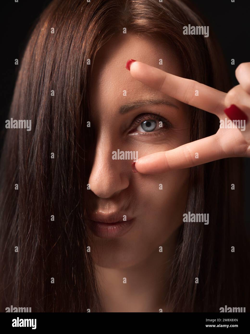 Gros plan portrait d'une femme montrant un geste de victoire ou de paix avec les doigts près de l'œil et regardant l'appareil photo. Vue de face de la dame brunette avec de longs cheveux Banque D'Images