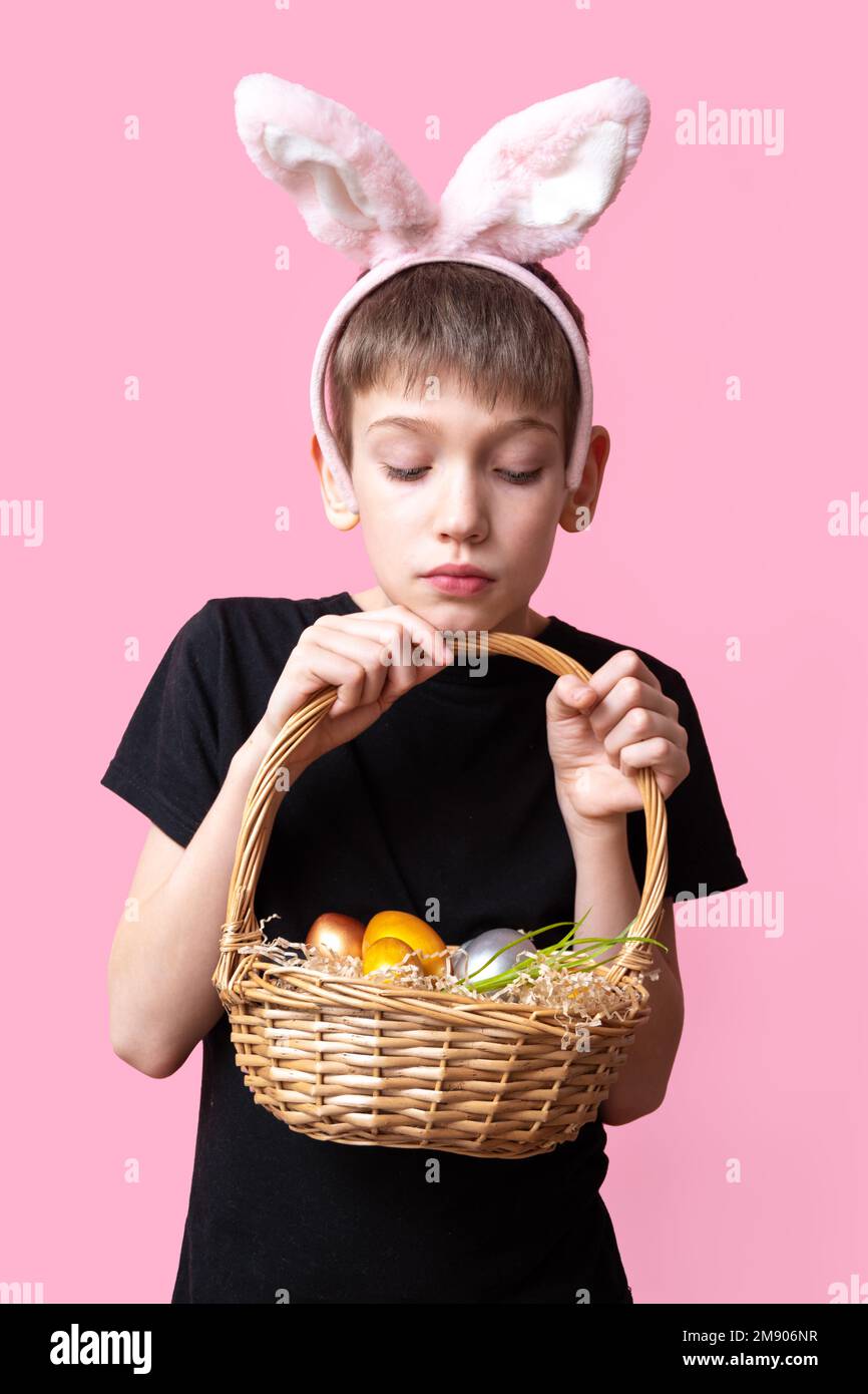 Un adolescent surpris avec des oreilles de lapin tient un panier avec des oeufs de Pâques colorés sur un fond rose, cadre vertical. Chasse aux œufs de lapin de Pâques. Banque D'Images