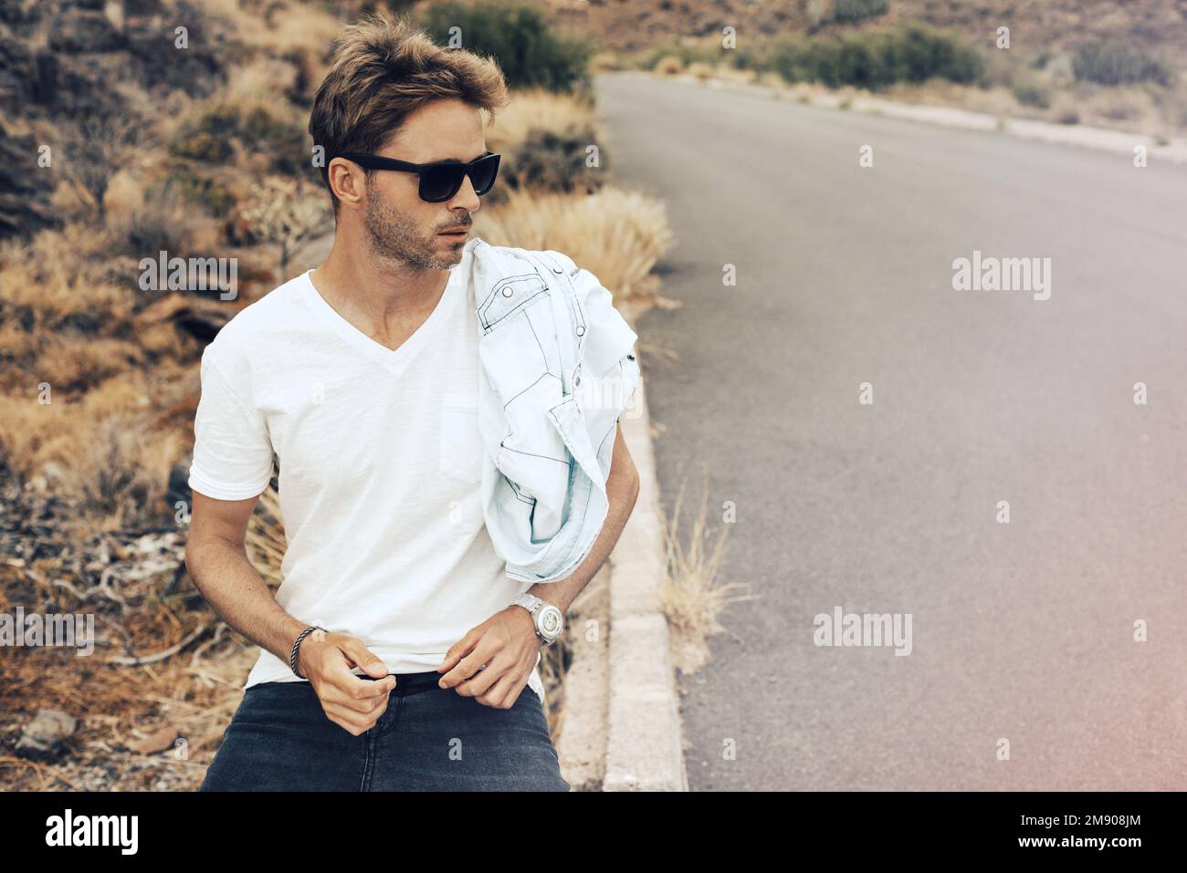 Jeune homme beau assis sur un parapet près de la colline rocheuse portant des lunettes, un t-shirt blanc et un Jean noir. Banque D'Images