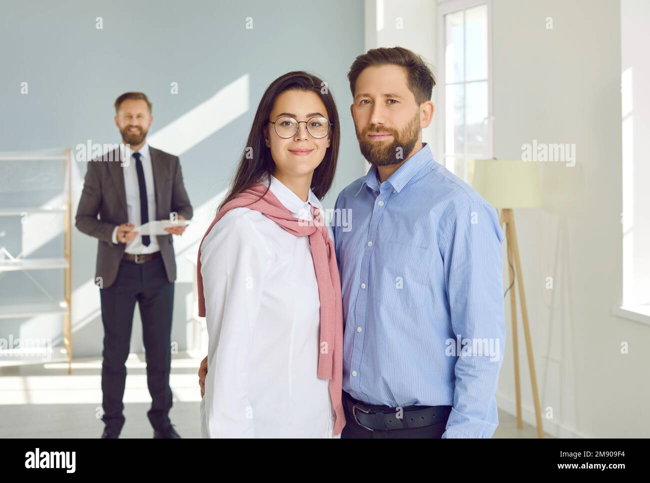 Portrait d'un jeune couple debout dans leur nouvelle maison, avec un agent immobilier en arrière-plan Banque D'Images