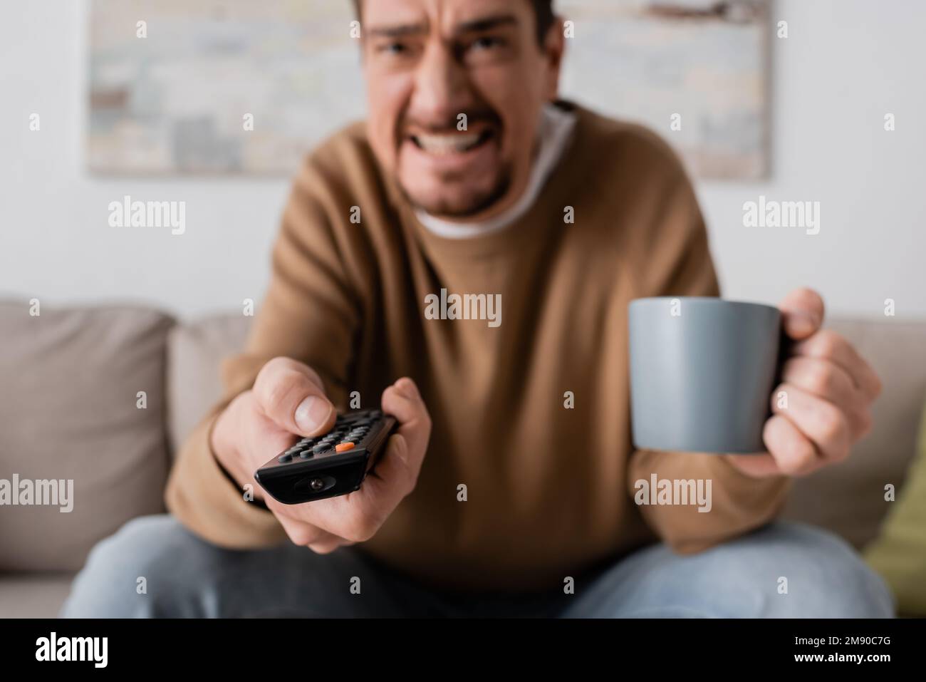 homme en colère tenant la télécommande et une tasse de café dans le salon, image de stock Banque D'Images